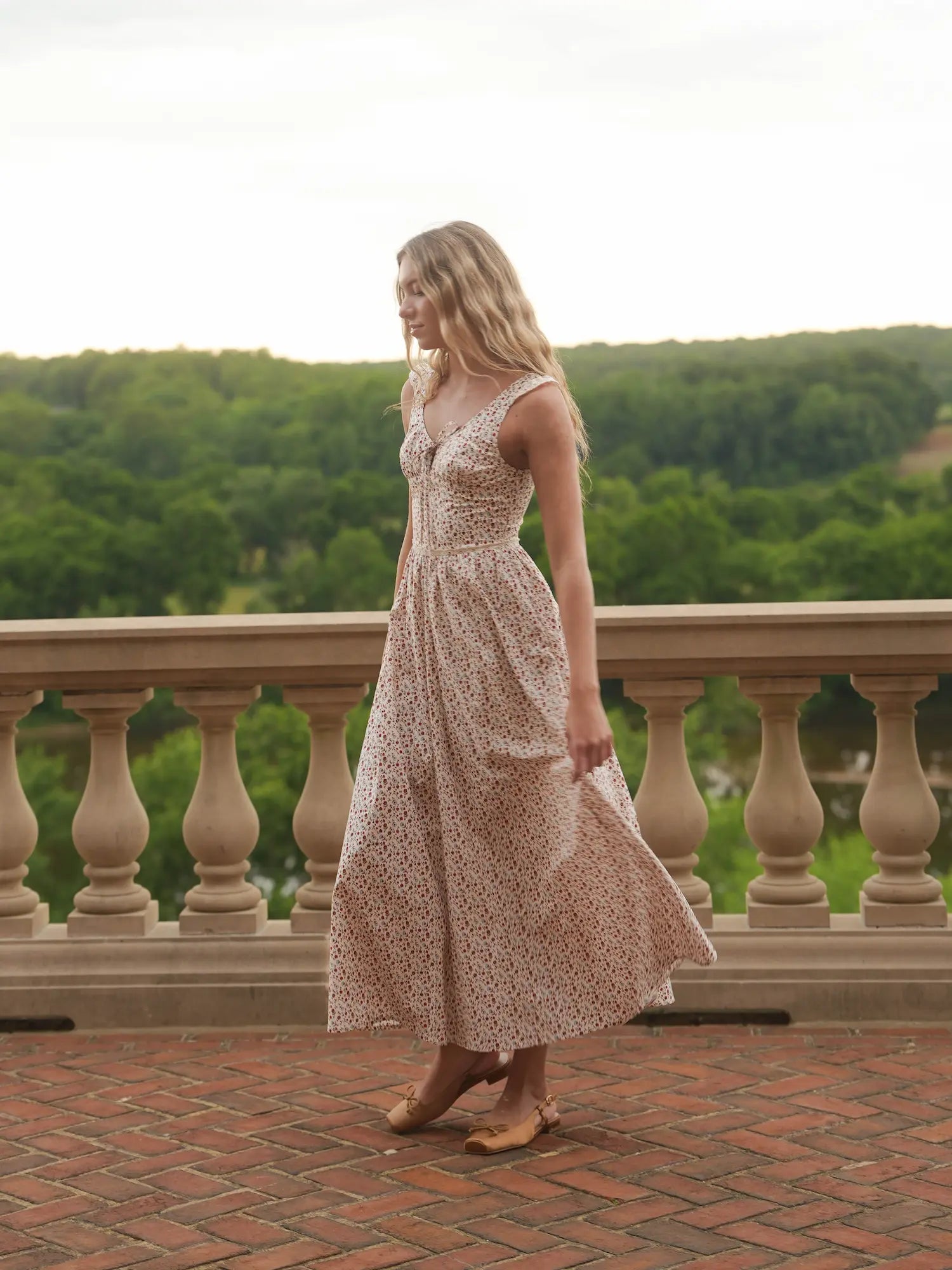 Woman in a floral dress standing on a balcony with a scenic view of trees.