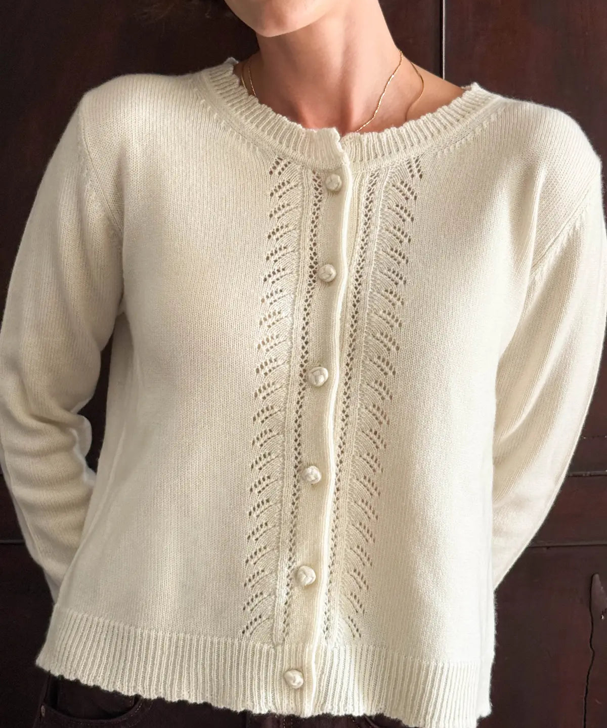A woman wearing a cream-colored knit cardigan sweater with decorative openwork patterns and a button-up front, shown against a dark wood background.