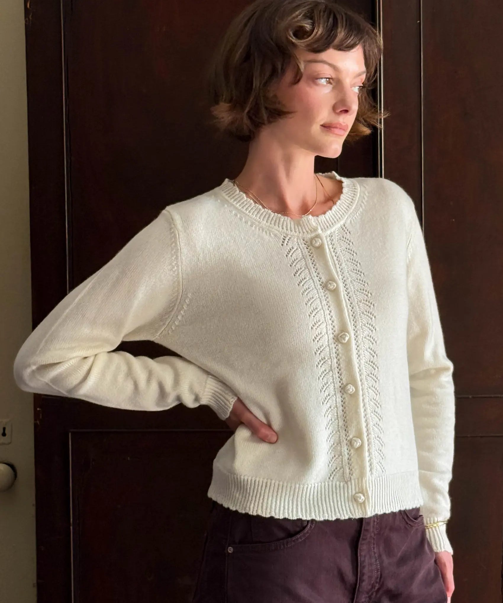 A woman wearing a cream-colored cable-knit cardigan sweater with frog buttons, paired with dark purple pants, standing in front of a dark wooden door.