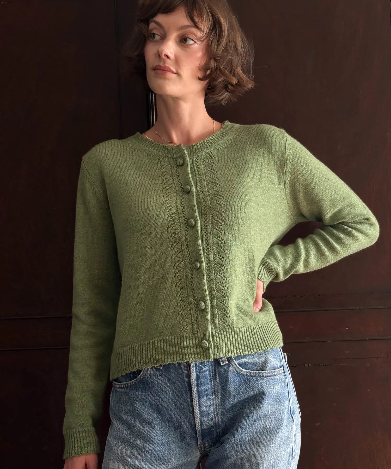 A woman wearing a light olive green knit cardigan sweater with button-front closure and blue jeans, standing against a dark brown wooden door.
