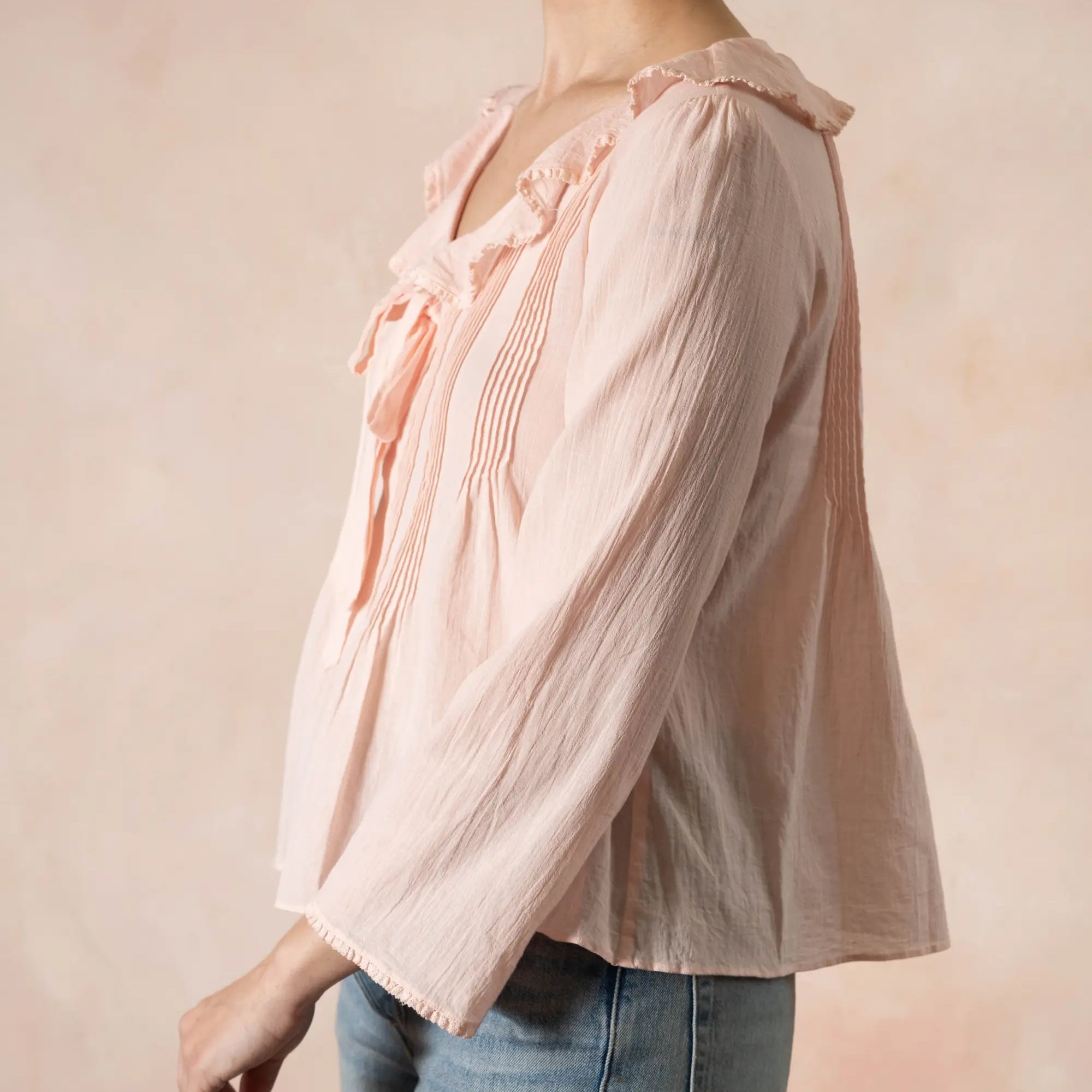 Person wearing a light pink blouse with ruffled details and blue jeans on a beige background