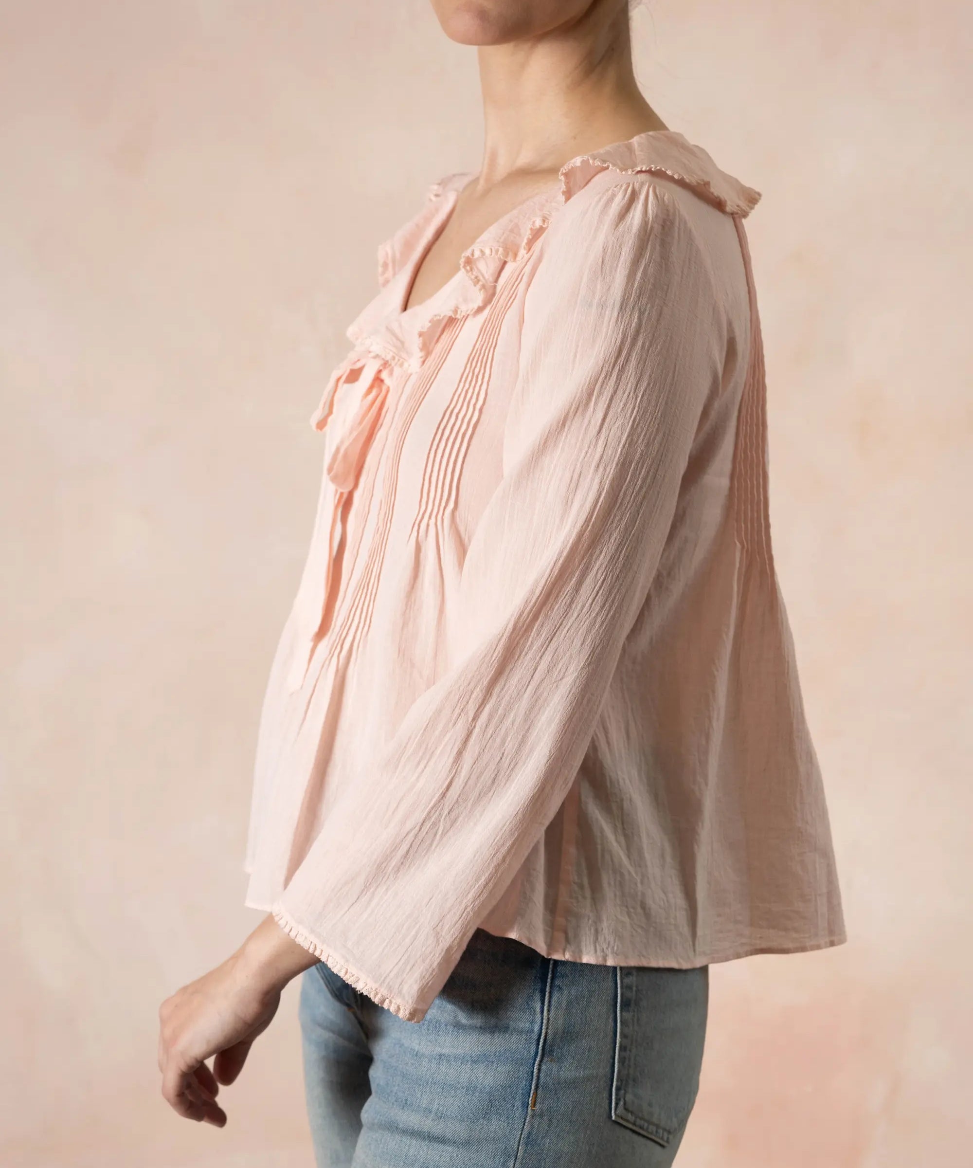 Person wearing a light pink blouse with ruffled details and blue jeans on a beige background