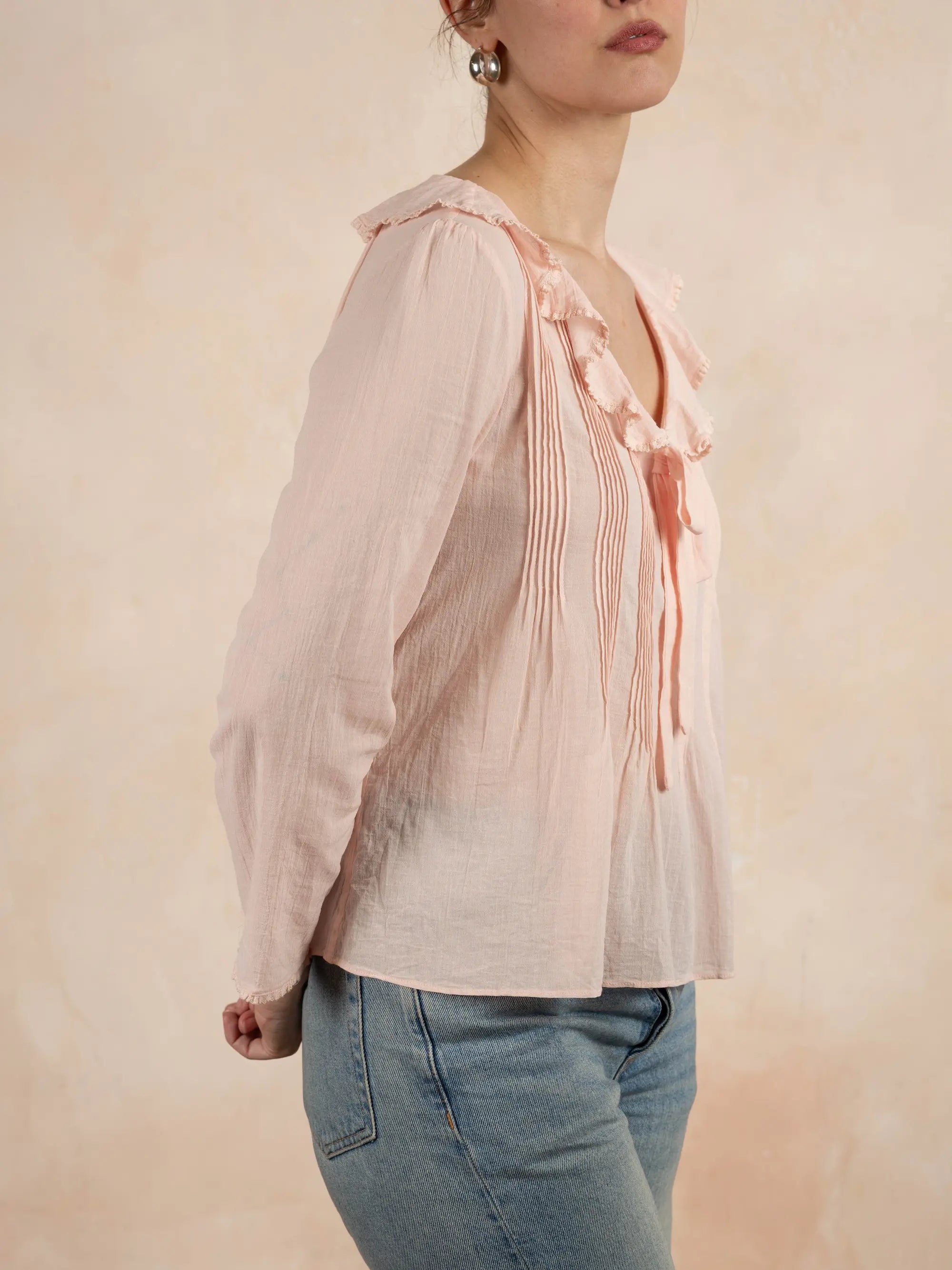 Person wearing a light pink blouse with ruffled details and blue jeans on a beige background