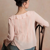 Woman wearing a light pink blouse sitting on a wooden chair against a beige wall.