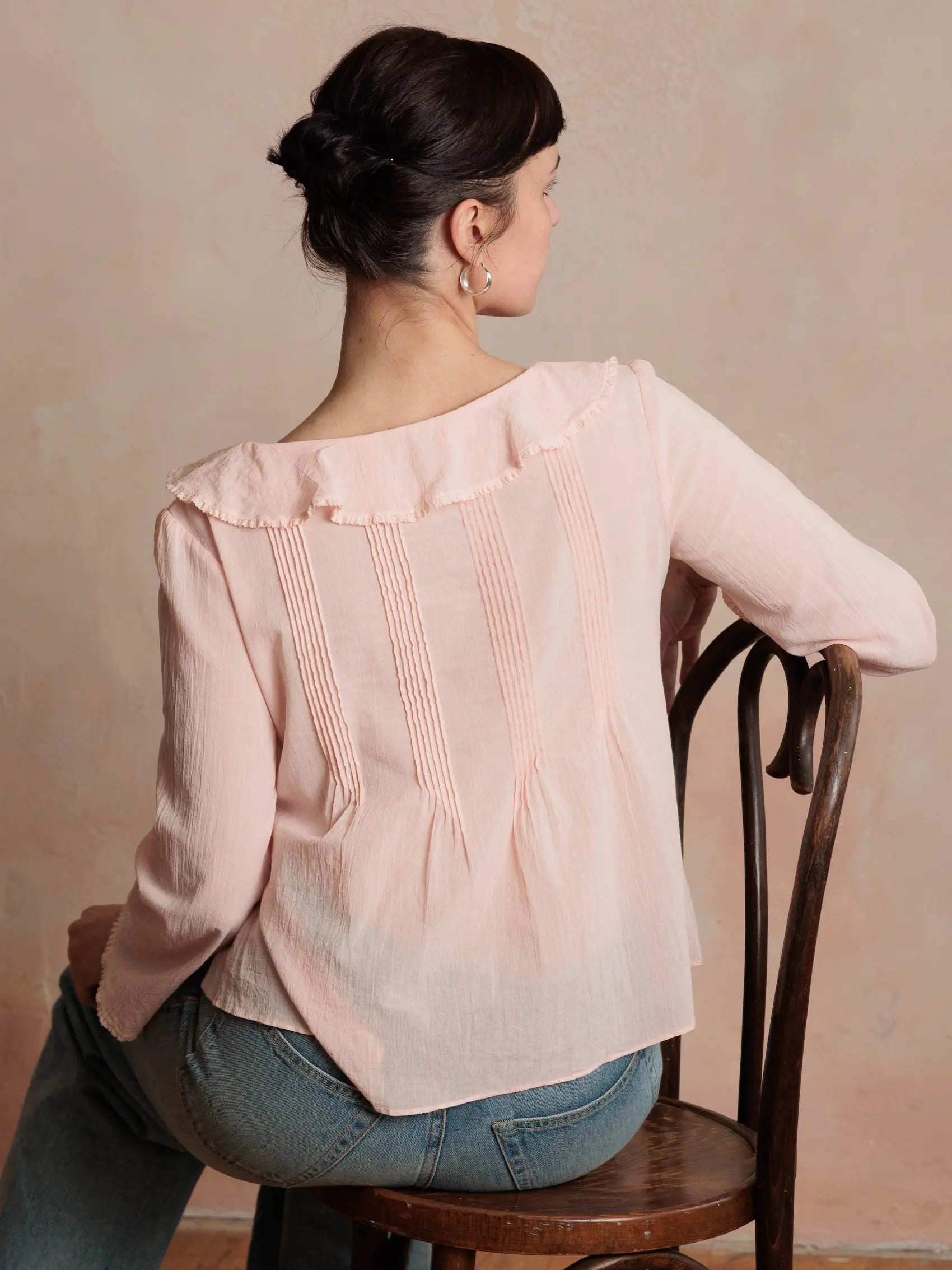 Woman wearing a light pink blouse sitting on a wooden chair against a beige wall.
