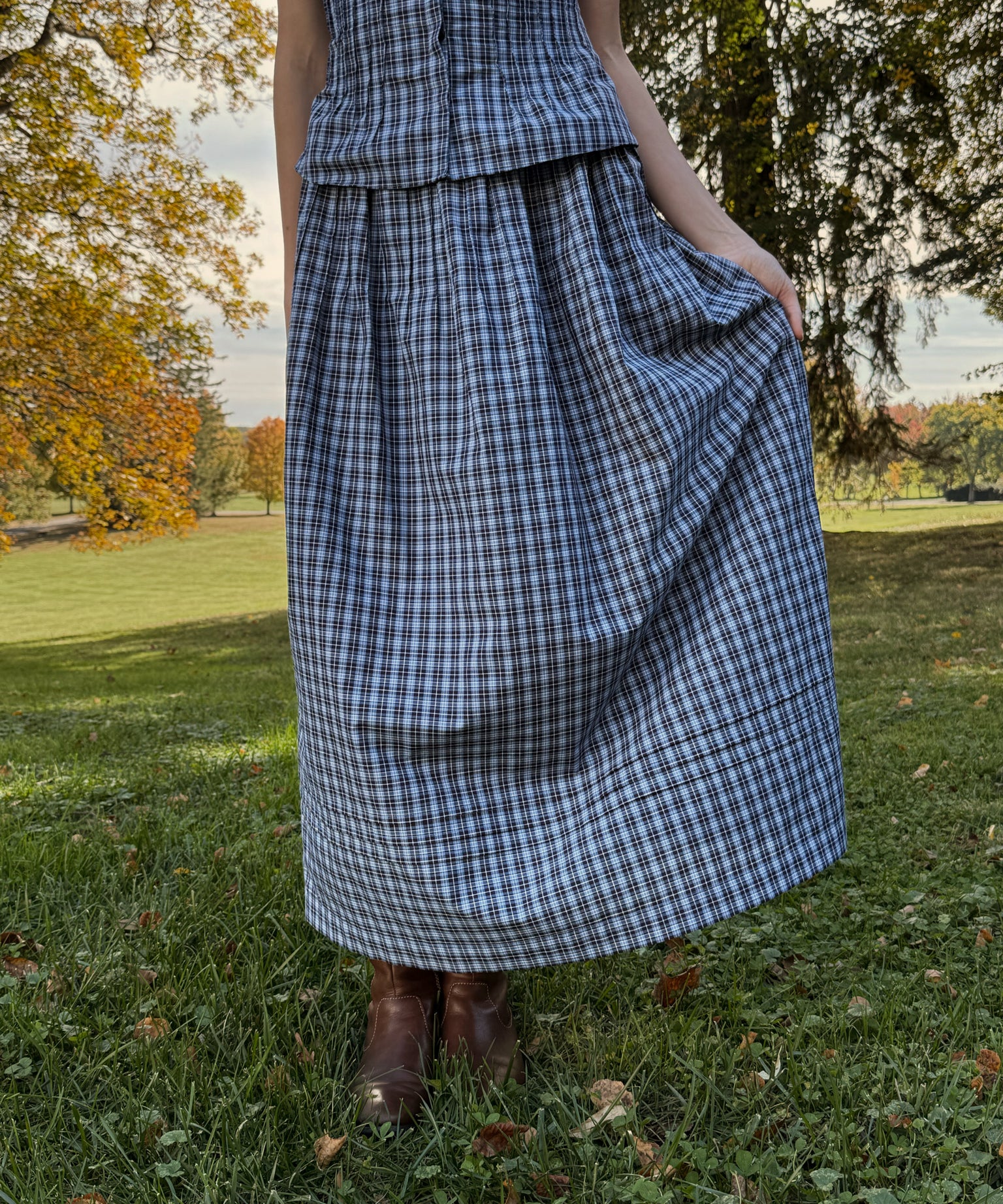 Person wearing a blue plaid skirt standing in a park with trees and grass.