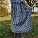 Person wearing a blue plaid dress standing in a park with trees and grass.