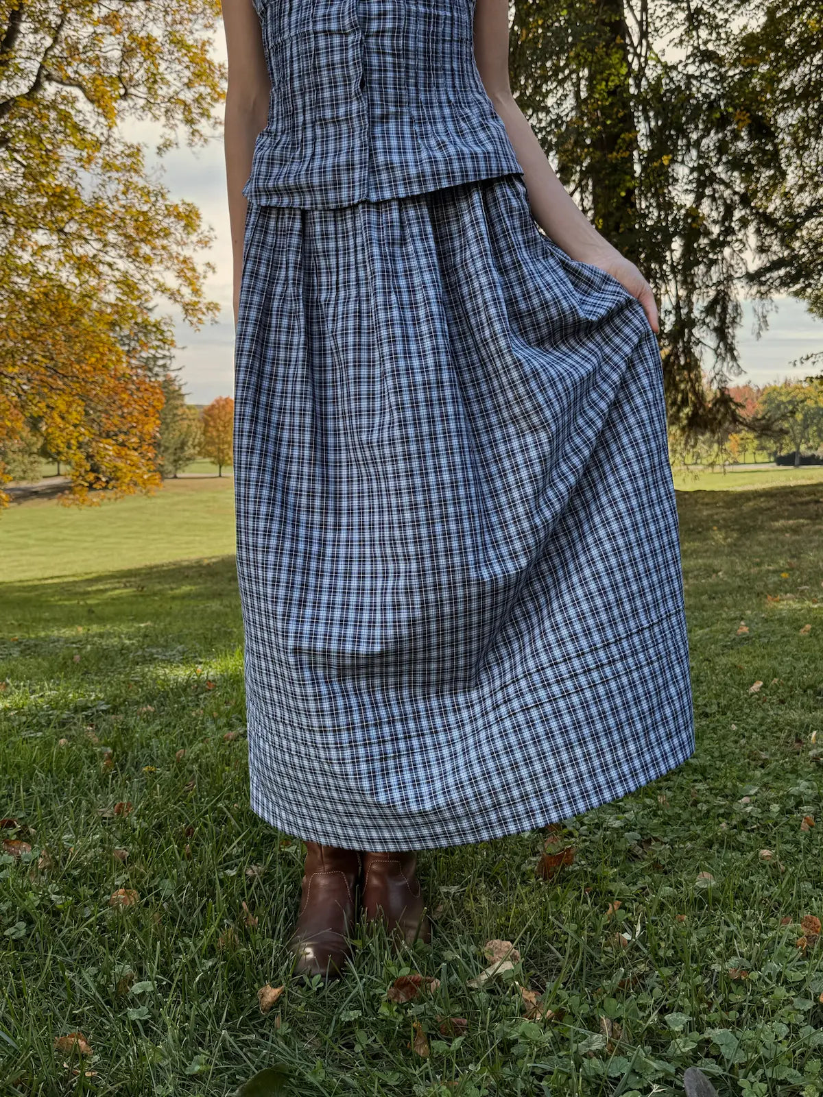 Person wearing a blue plaid dress standing in a park with trees and grass.