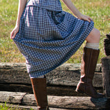 Person wearing a blue plaid set and brown boots standing on wooden planks in a grassy field.