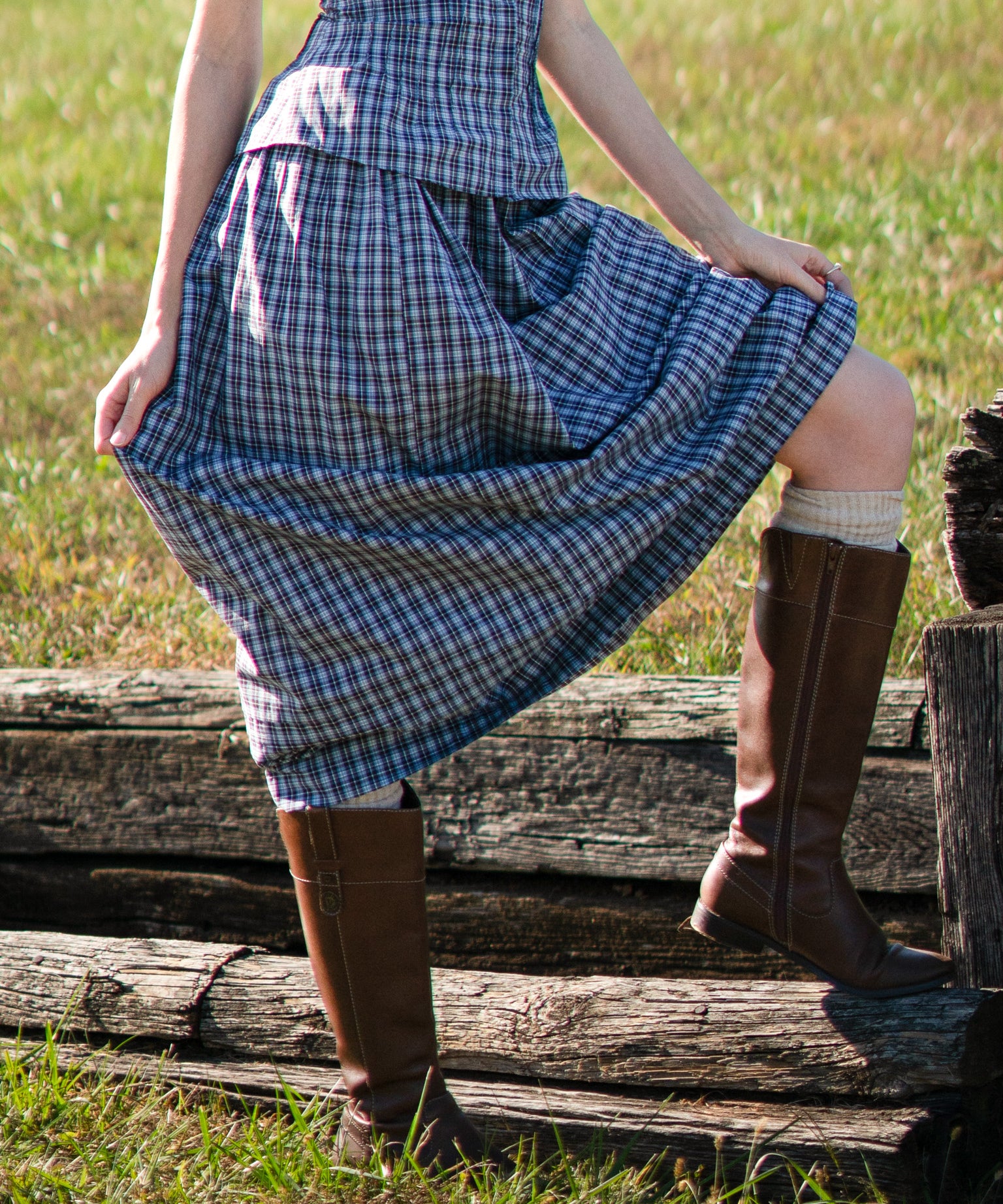 Person wearing a blue plaid set and brown boots standing on wooden planks in a grassy field.
