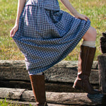 Person wearing a blue plaid dress and brown boots standing on wooden planks in a grassy field.