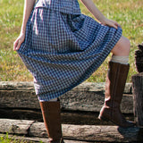 Person wearing a blue plaid dress and brown boots standing on wooden planks in a grassy field.