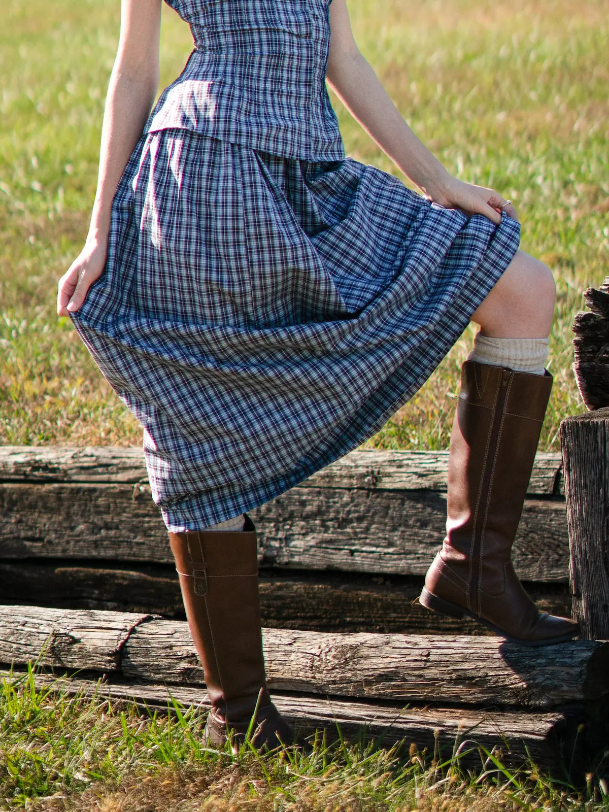 Person wearing a blue plaid dress and brown boots standing on wooden planks in a grassy field.