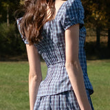 Woman wearing a blue plaid dress standing in a grassy area with trees in the background