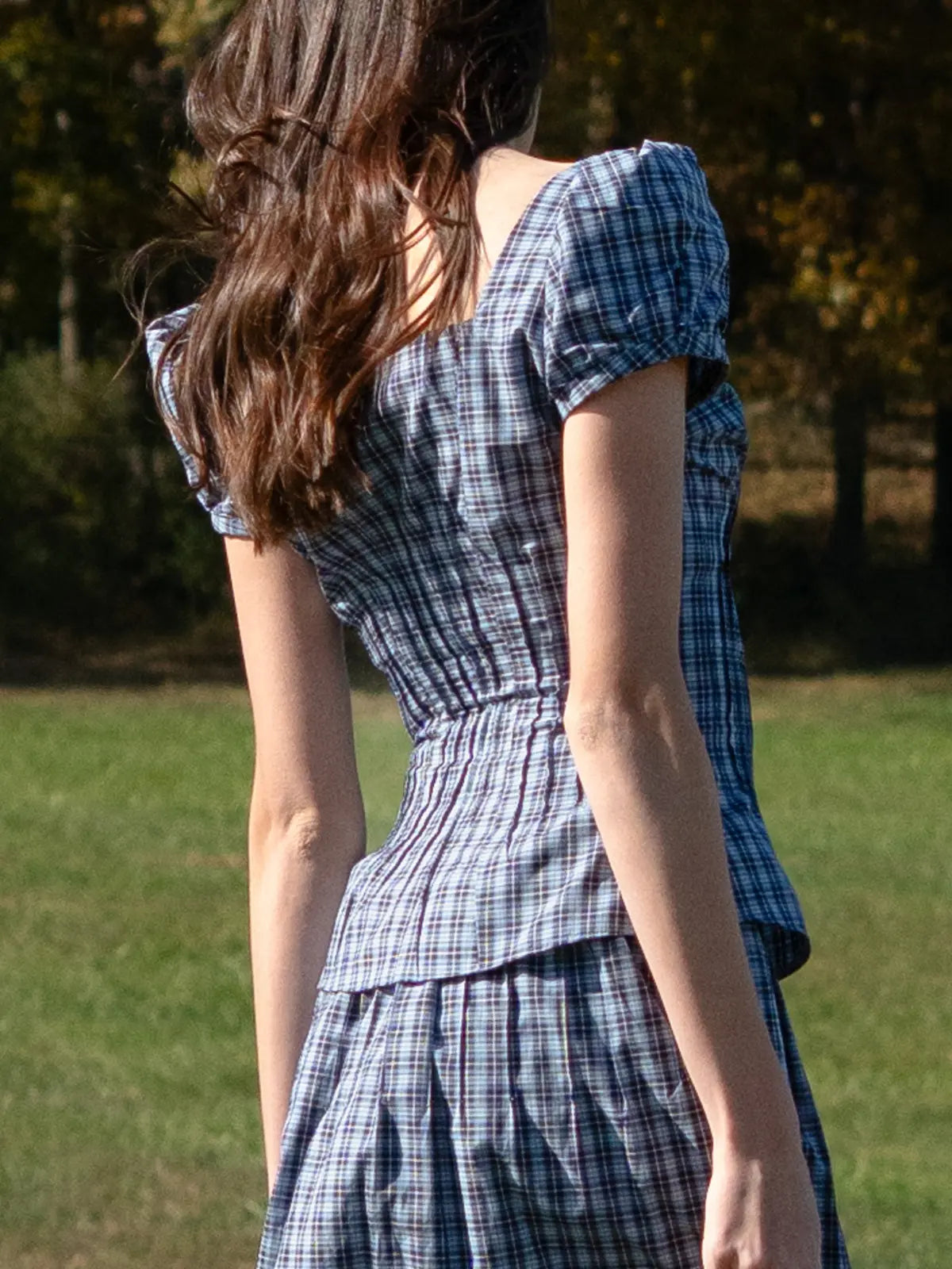 Woman wearing a blue plaid dress standing in a grassy area with trees in the background