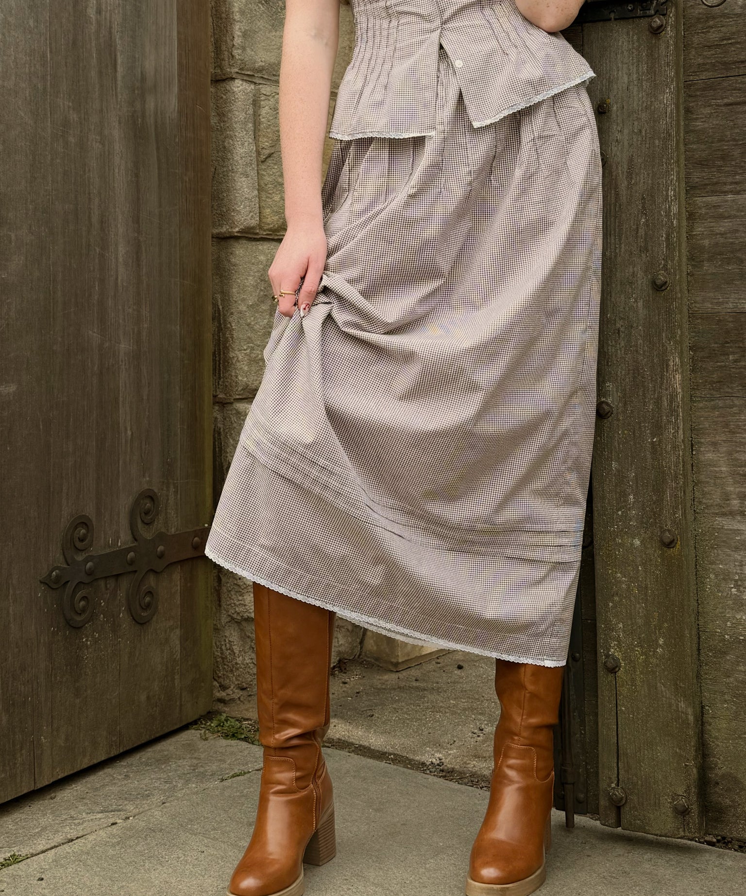 Woman wearing a brown gingham print skirt and brown boots standing against a wooden door.
