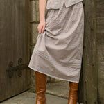 Person wearing a long brown gingham skirt and brown boots standing in front of a wooden door.