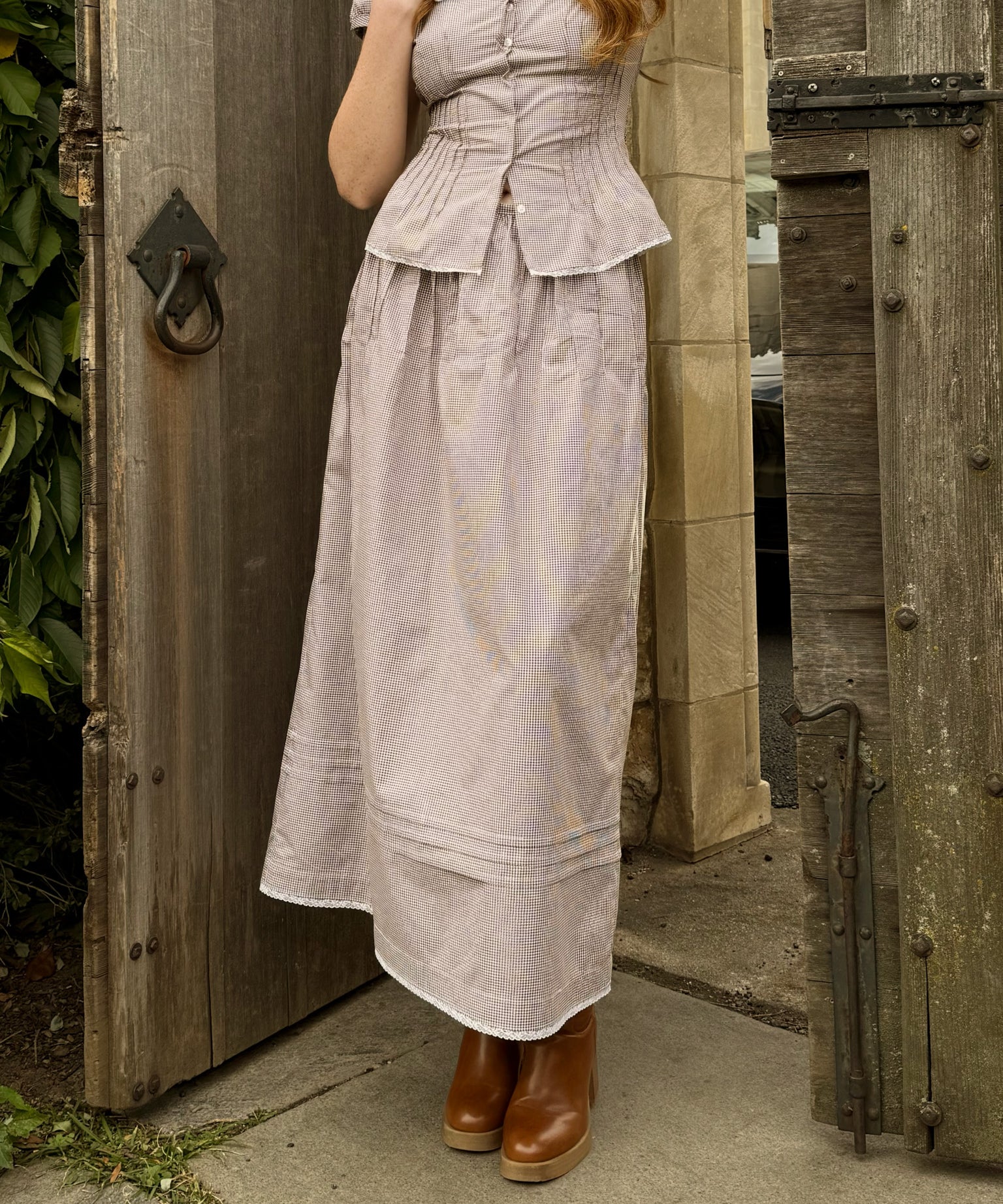 Woman wearing a brown gingham print skirt dress with a matching peplum top, standing in front of a wooden door.