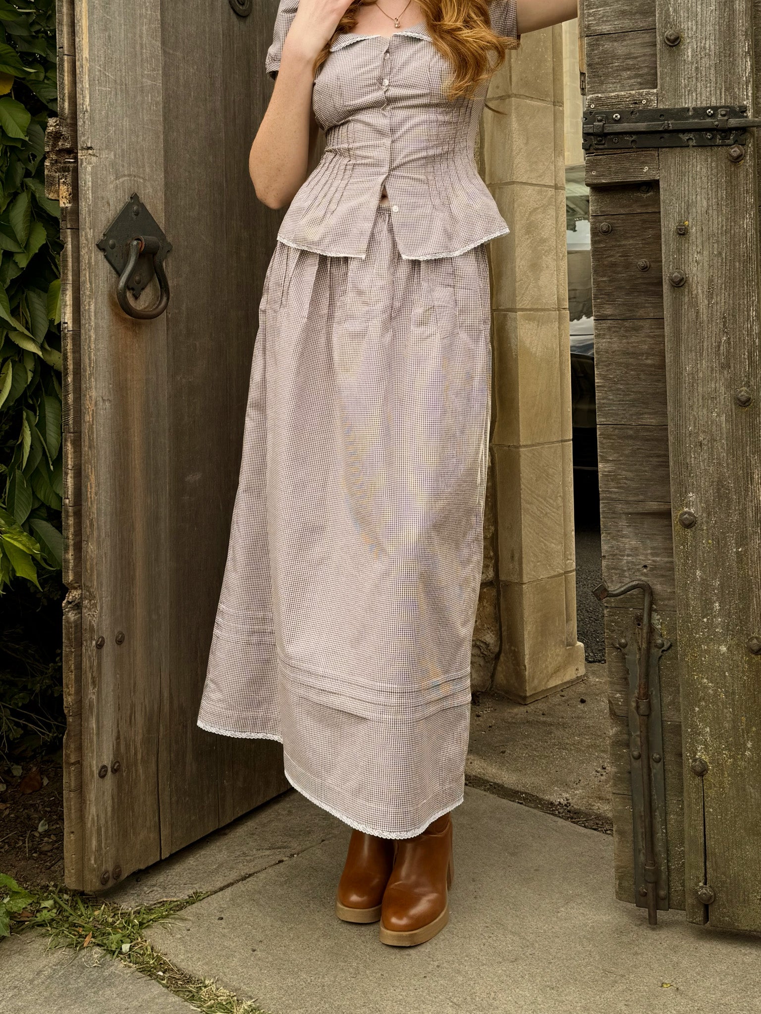 Woman wearing a brown gingham print skirt dress with a matching peplum top, standing in front of a wooden door.