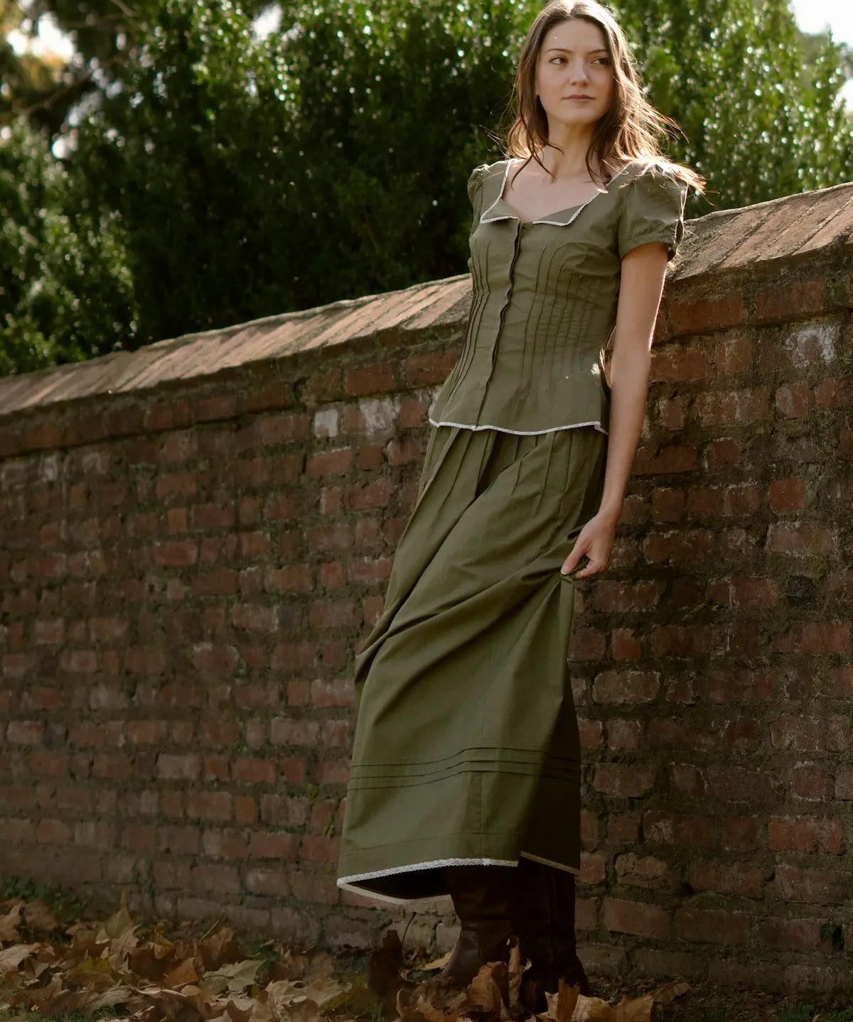 Woman in a green dress leaning against a brick wall with trees in the background