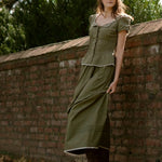Woman in a green dress leaning against a brick wall with trees in the background
