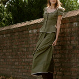 Woman in a green dress leaning against a brick wall with trees in the background