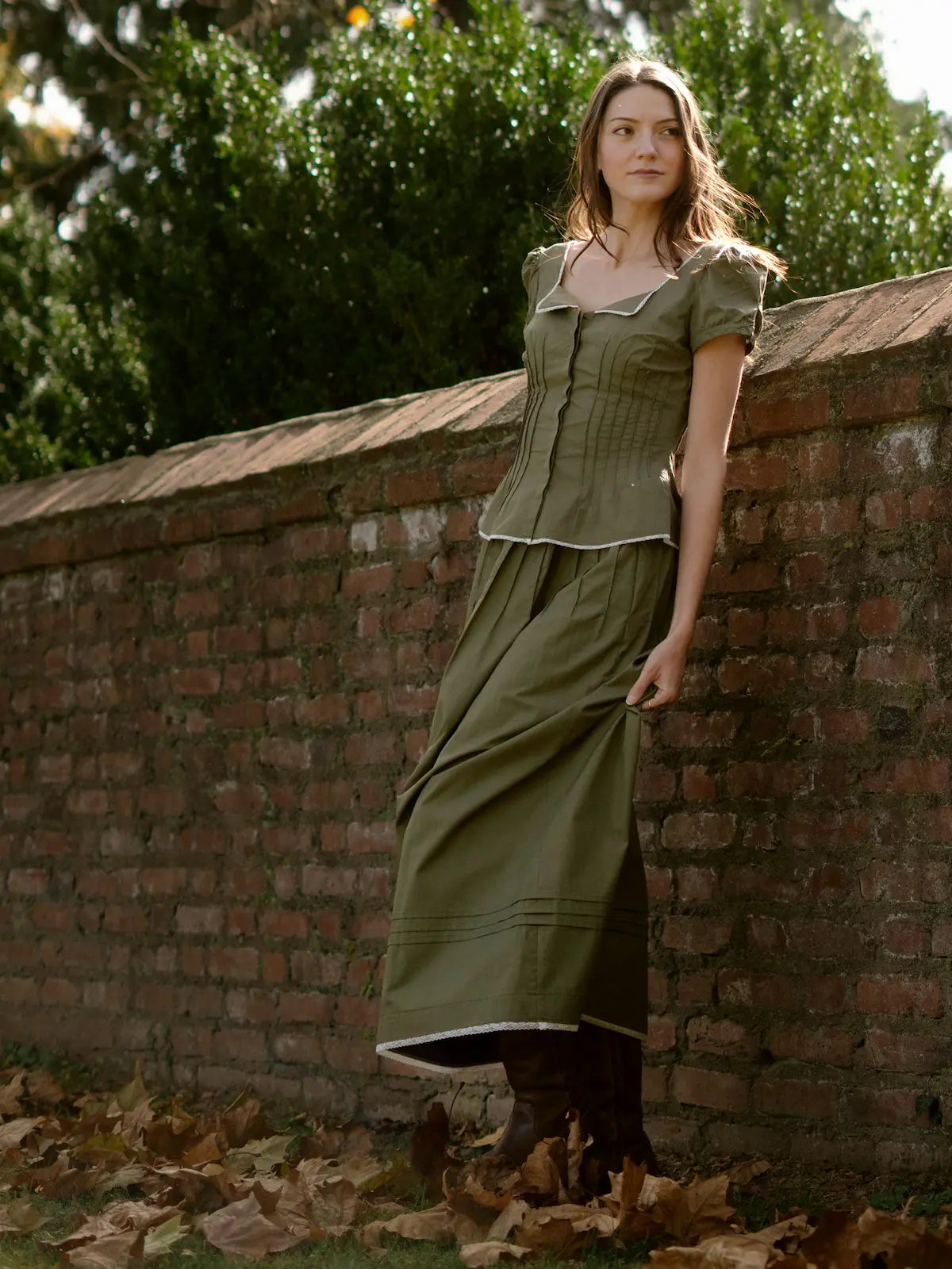 Woman in a green dress leaning against a brick wall with trees in the background