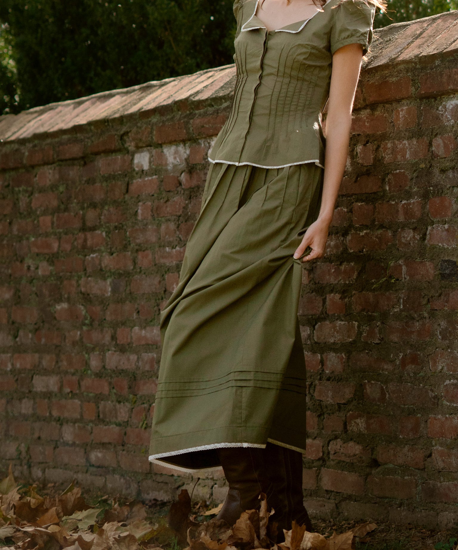 Person wearing a green set leaning against a brick wall with fallen leaves on the ground.