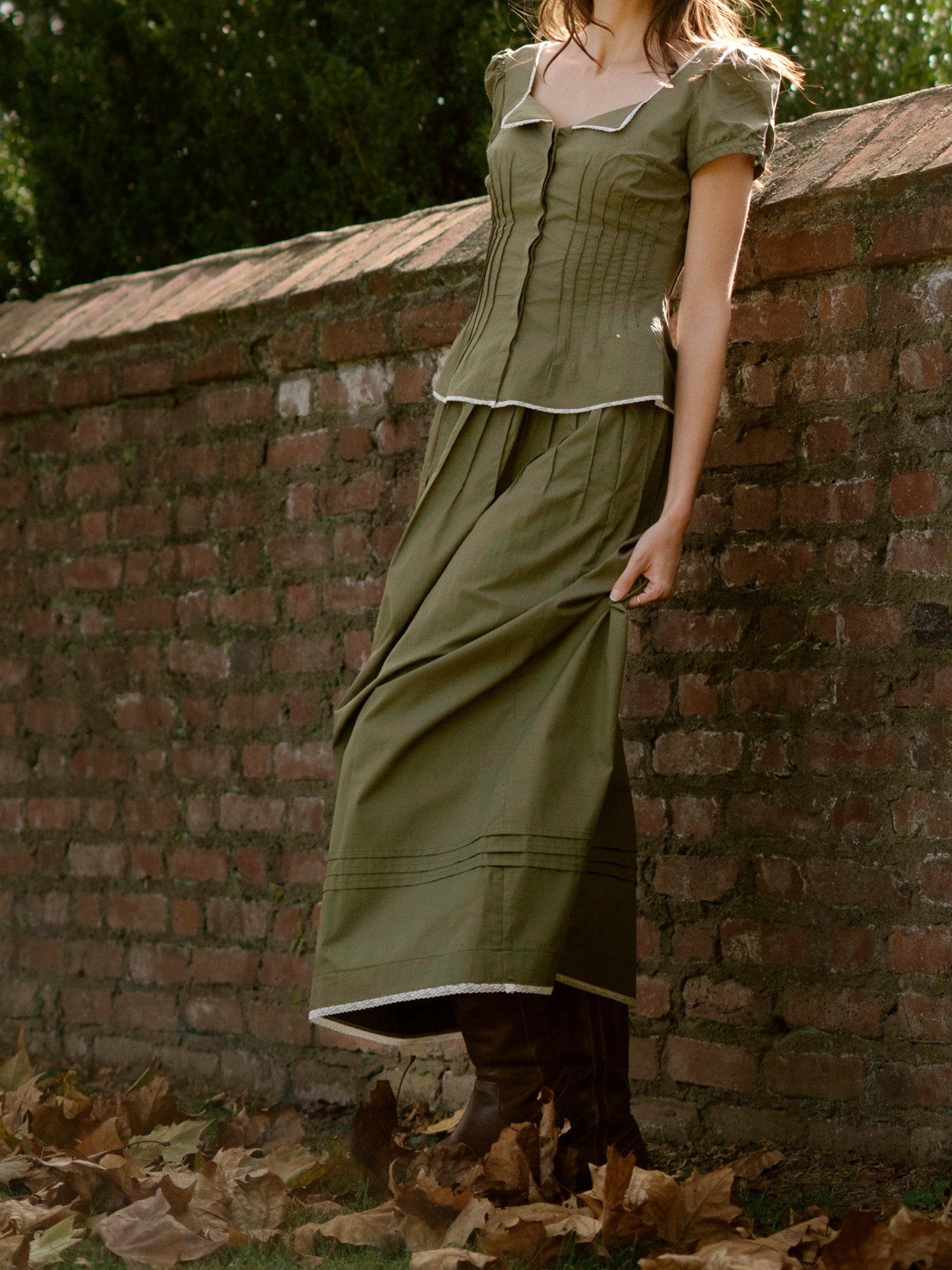 Person wearing a green set leaning against a brick wall with fallen leaves on the ground.