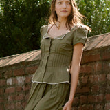 Woman in an olive green top and skirt leaning against a brick wall with greenery in the background