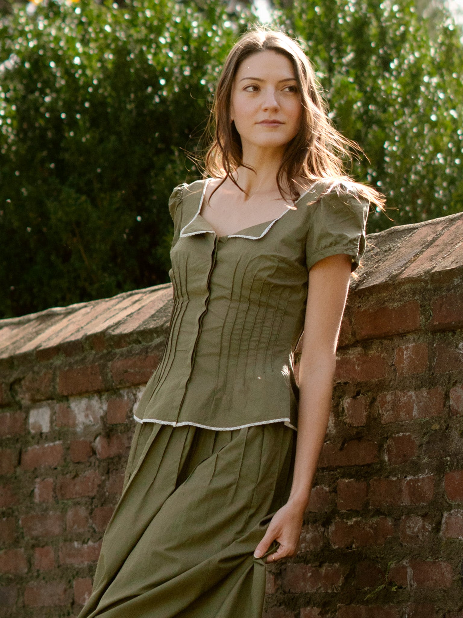 Woman in an olive green top and skirt leaning against a brick wall with greenery in the background