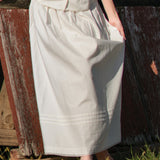 Person wearing a long light-colored skirt standing next to a wooden fence.